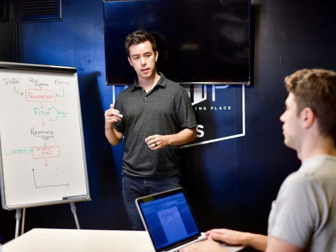 Ein Mann steht vor einen Flipchart. Das Flipchart ist beschriftet. Ein weiterer Mann schreibt an seinem Laptop.